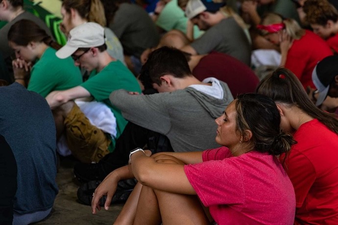 A group of people sit closely together on the ground with heads bowed and eyes closed, appearing to be in prayer or deep thought. The atmosphere is quiet and reflective.