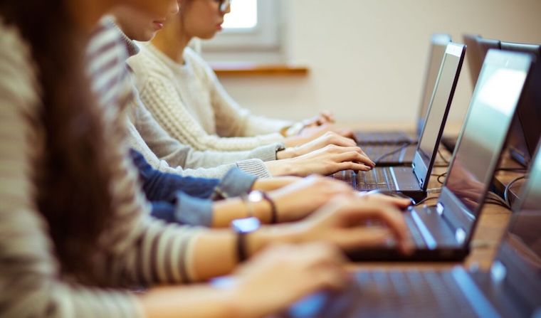 A row of students sitting at desks, working on laptop computers in a classroom setting. The focus is on their hands typing, with faces partially visible and blurred.