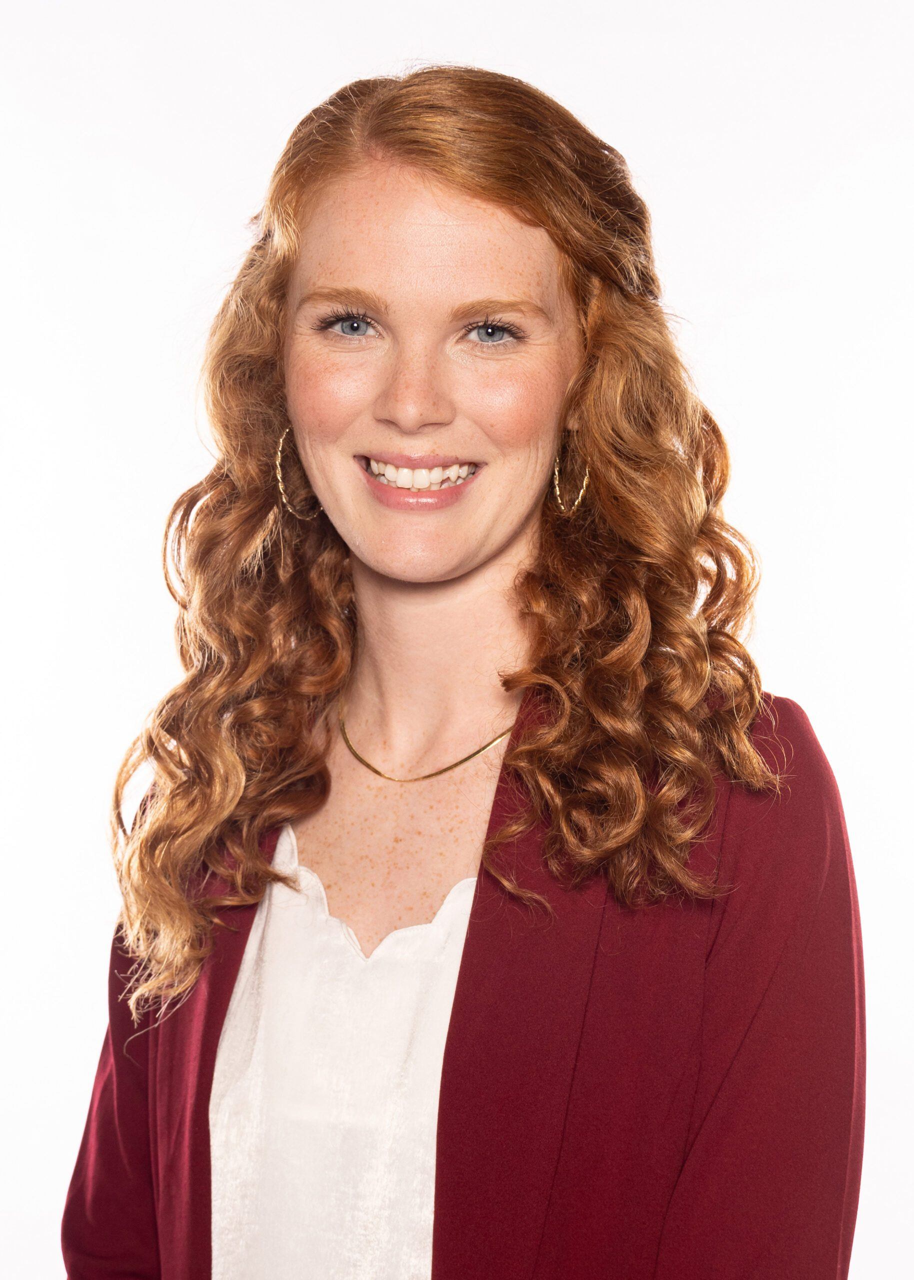 A woman with long, curly red hair smiles at the camera. She is wearing a maroon blazer over a white blouse, gold hoop earrings, and a simple gold necklace. The background is plain white.