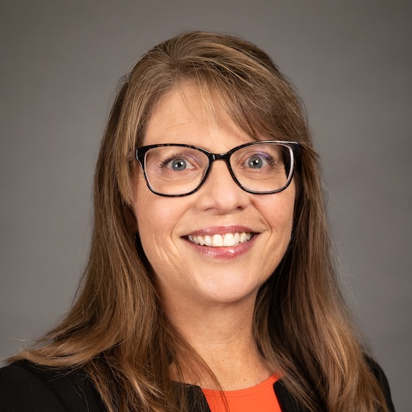 Smiling woman with light brown hair in a bun, wearing black-rimmed glasses, hoop earrings, a dark blazer, and a floral top, posed against a plain dark background.