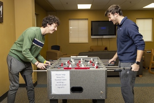 Two young men smiling and playing foosball in a lounge area. A sign on the table reads, Please help keep this foosball table working. Do not sit on top of it. Sofas and a TV are visible in the background.