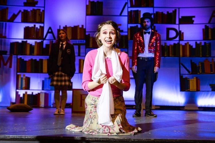 A young woman kneels center stage under bright lights, wearing a pink sweater and floral skirt, smiling emotionally. Two people stand behind her against a colorful backdrop of bookshelves and large letters.