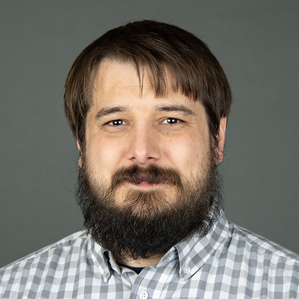 A man with a dark beard and mustache, wearing a light gray and white checkered shirt, poses in front of a plain gray background.