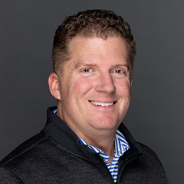 A man with short curly brown hair smiles at the camera. He is wearing a dark jacket over a blue and white striped collared shirt. The background is plain and dark gray.