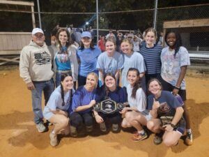 A softball team poses on a dirt field at night, smiling for a group photo. One player in front holds a championship-style belt. Players and two adults stand together, with a fence and spectators in the background.