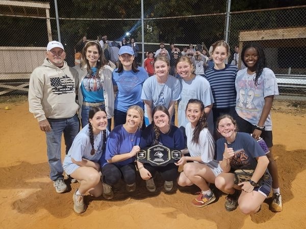 A softball team poses on a dirt field at night, smiling for a group photo. One player in front holds a championship-style belt. Players and two adults stand together, with a fence and spectators in the background.
