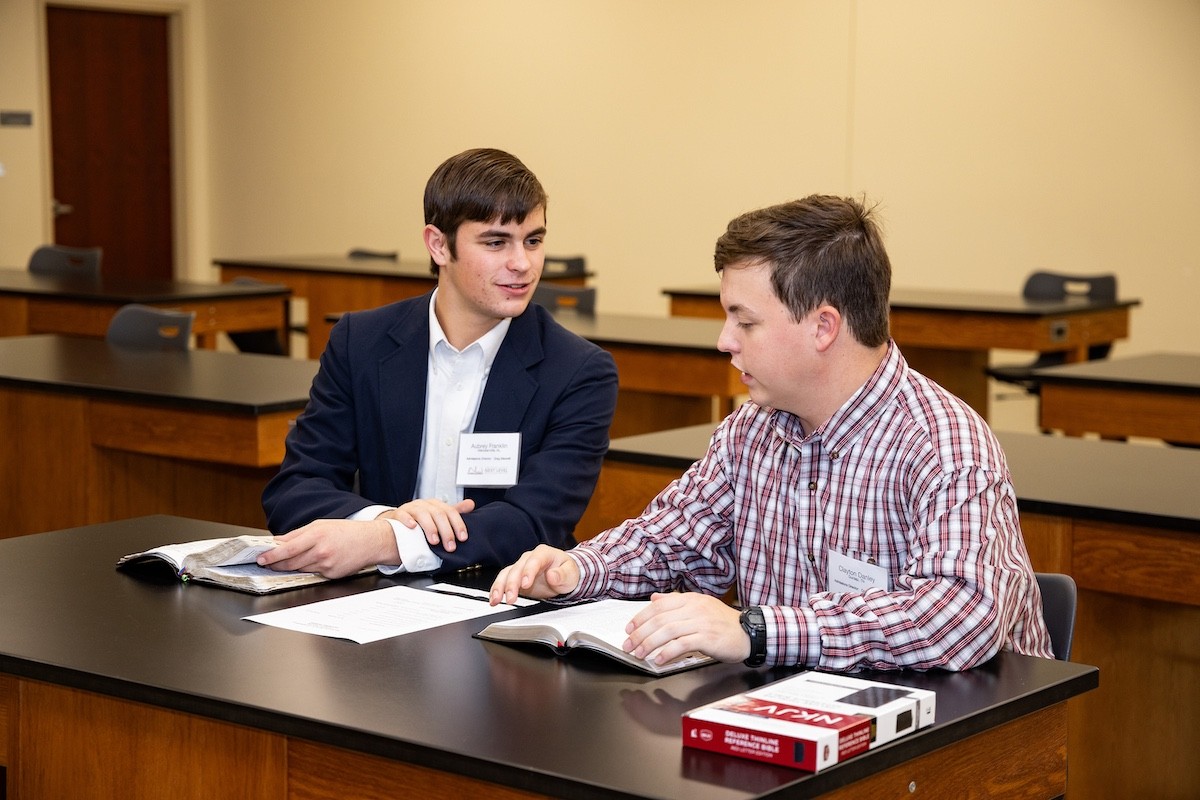 Two young men sit at a classroom table, engaged in conversation. Both have open books in front of them and wear name tags. The classroom has empty wooden tables and a plain beige wall.